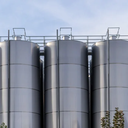 Three large, cylindrical metal storage silos, often used by an Edible Oil Refinery Plant Manufacturer, are lined up outdoors against a blue sky, with a few tree branches visible at the bottom right.